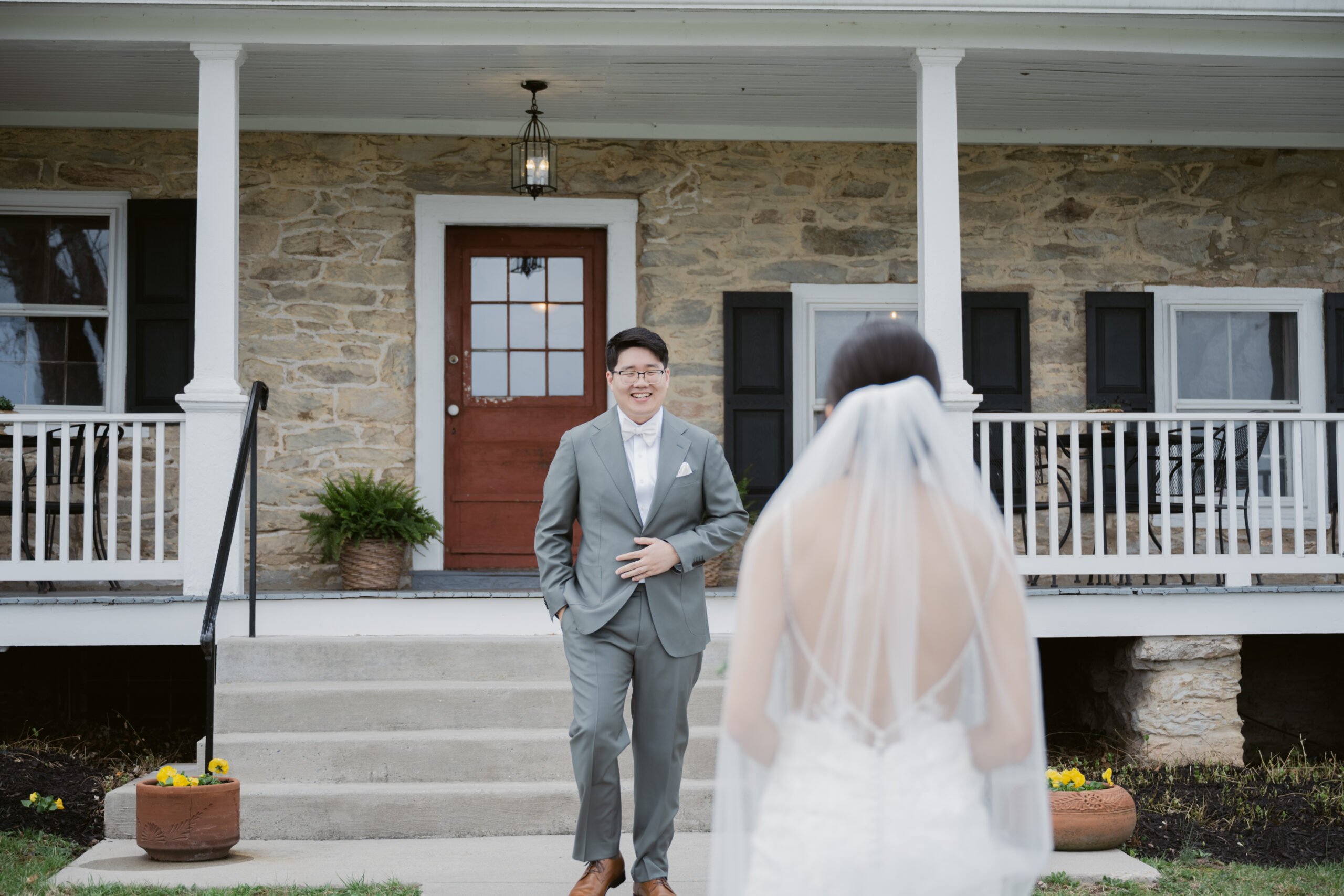 Groom smiling as he sees his bride for the first time during their first look outside a stone farmhouse at Springfield Manor, with the bride approaching in her wedding gown and veil.