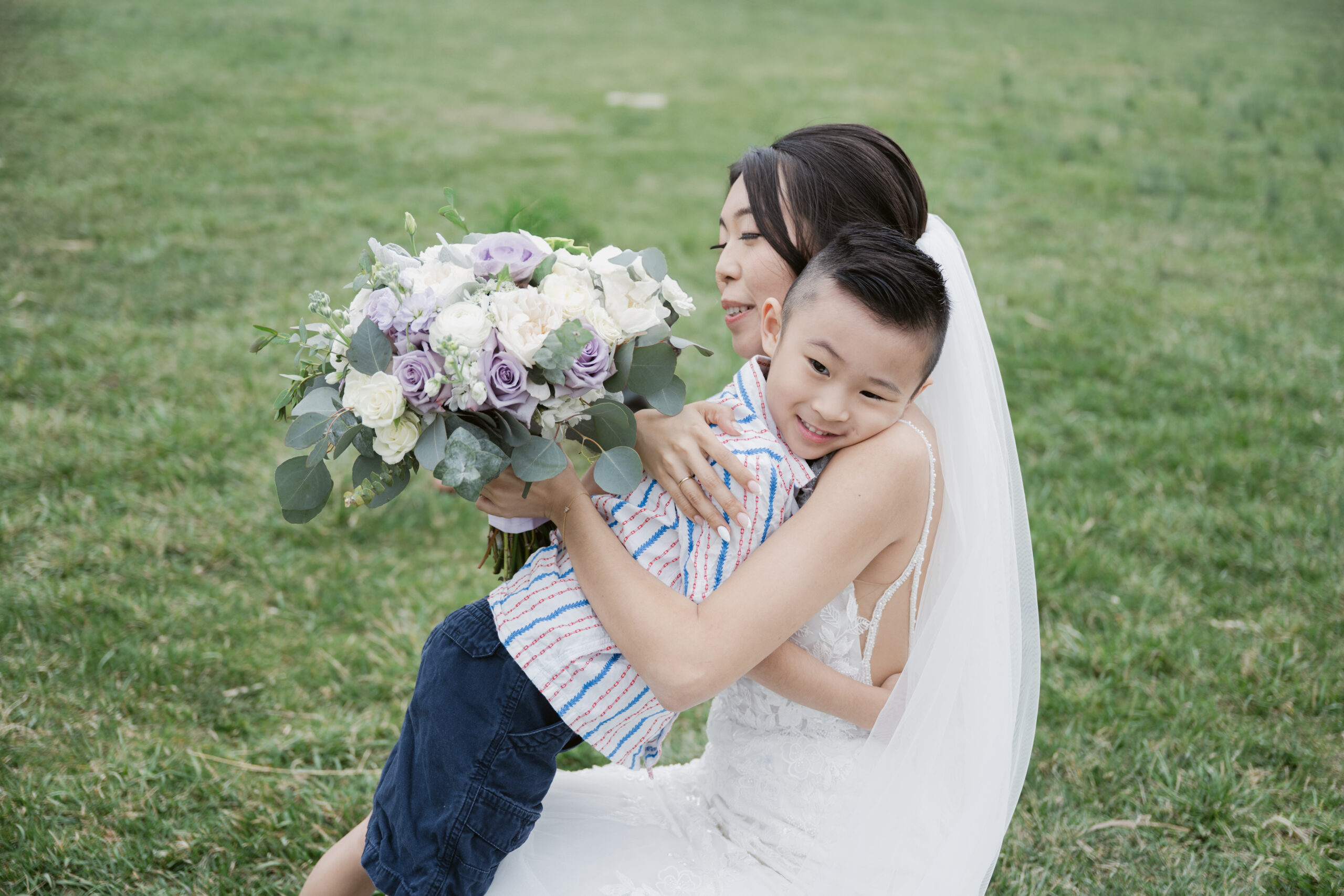 Bride crouching to hug a smiling young boy while holding a bouquet of white and purple flowers.