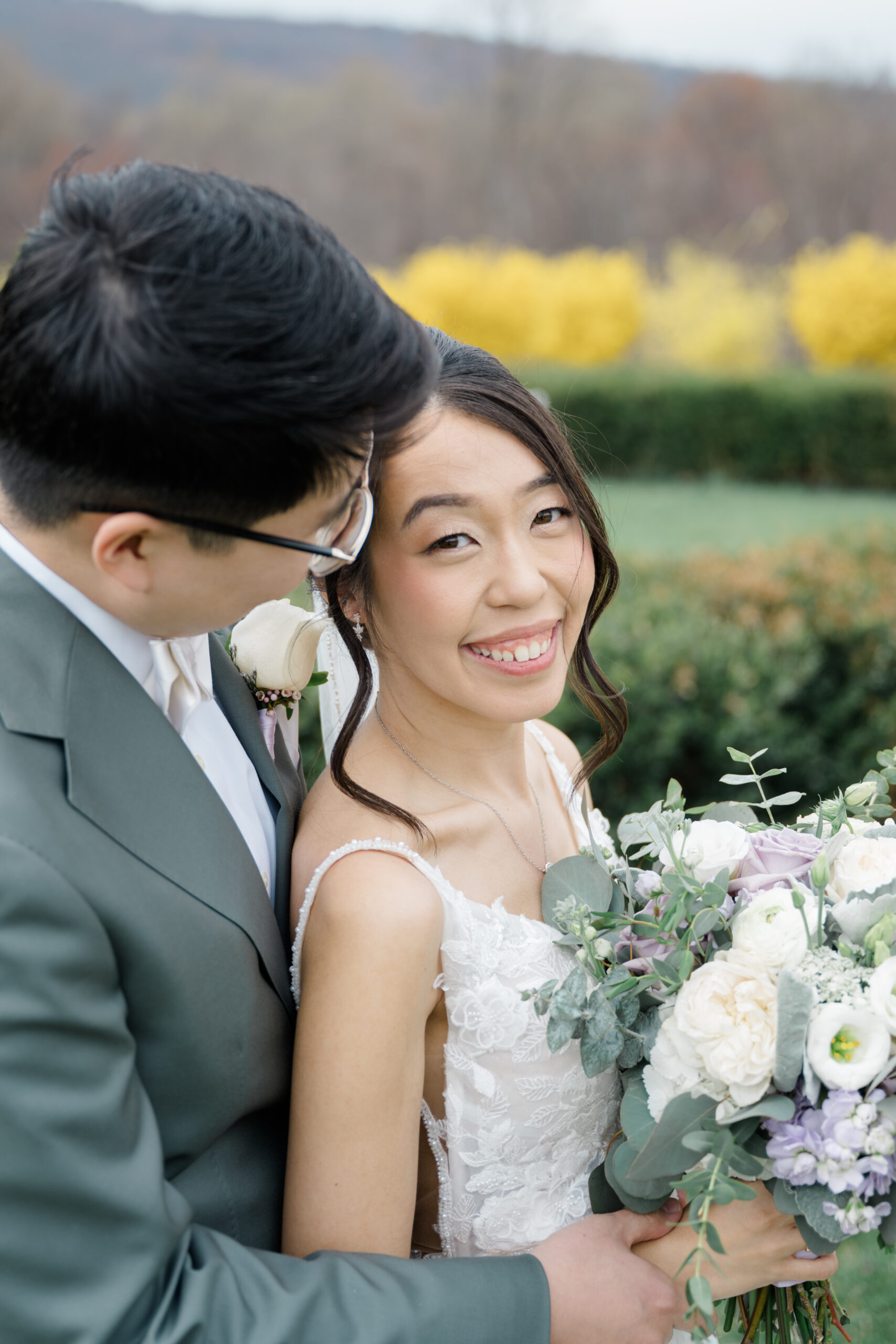 Groom smiling down at bride as she looks at the camera while holding a bouquet of white and lavender flowers.