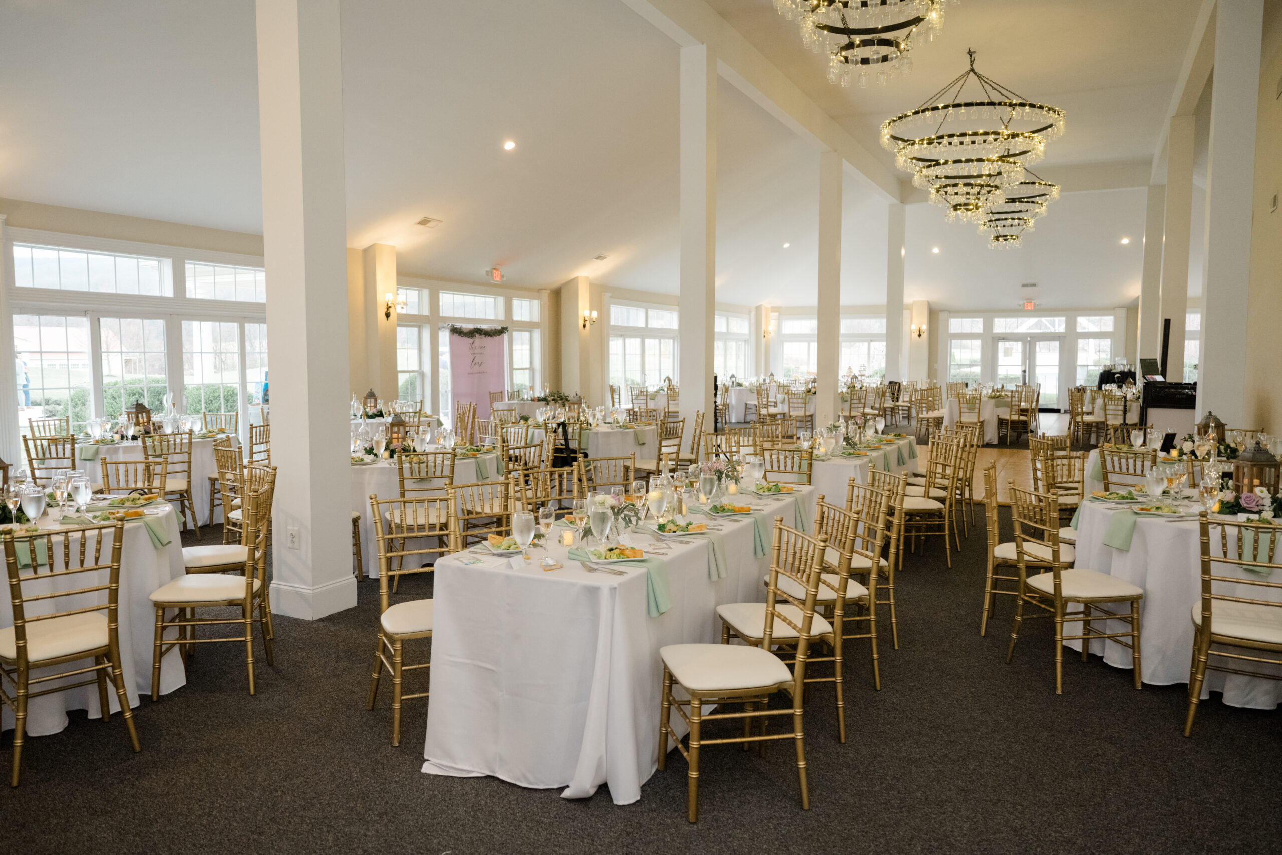 Reception hall setup with gold chairs, white tablecloths, and elegant table settings under chandeliers.
