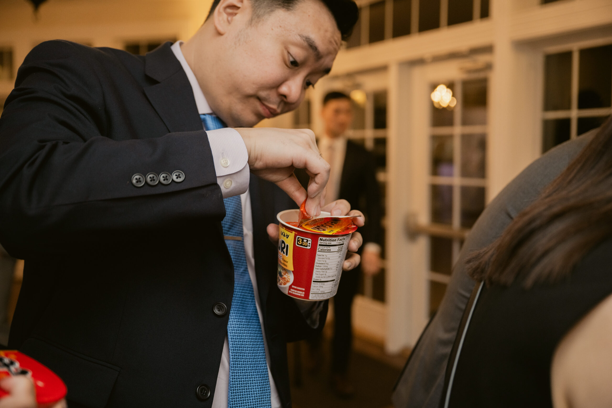 Wedding guest in a suit opening a cup of instant ramen with focused expression.