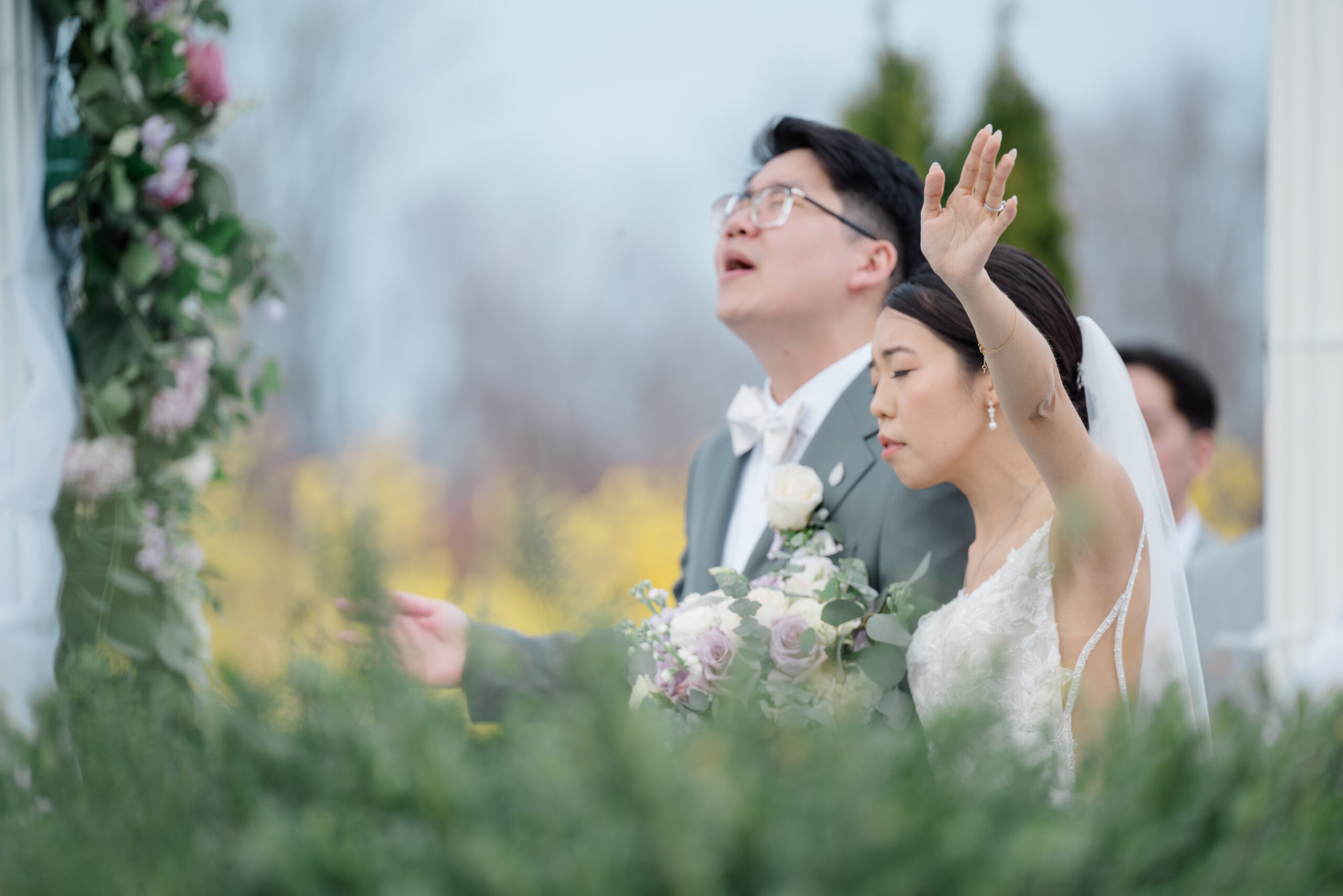 Bride and groom worship with eyes closed and hands raised during their outdoor wedding ceremony, surrounded by soft greenery and spring florals.
