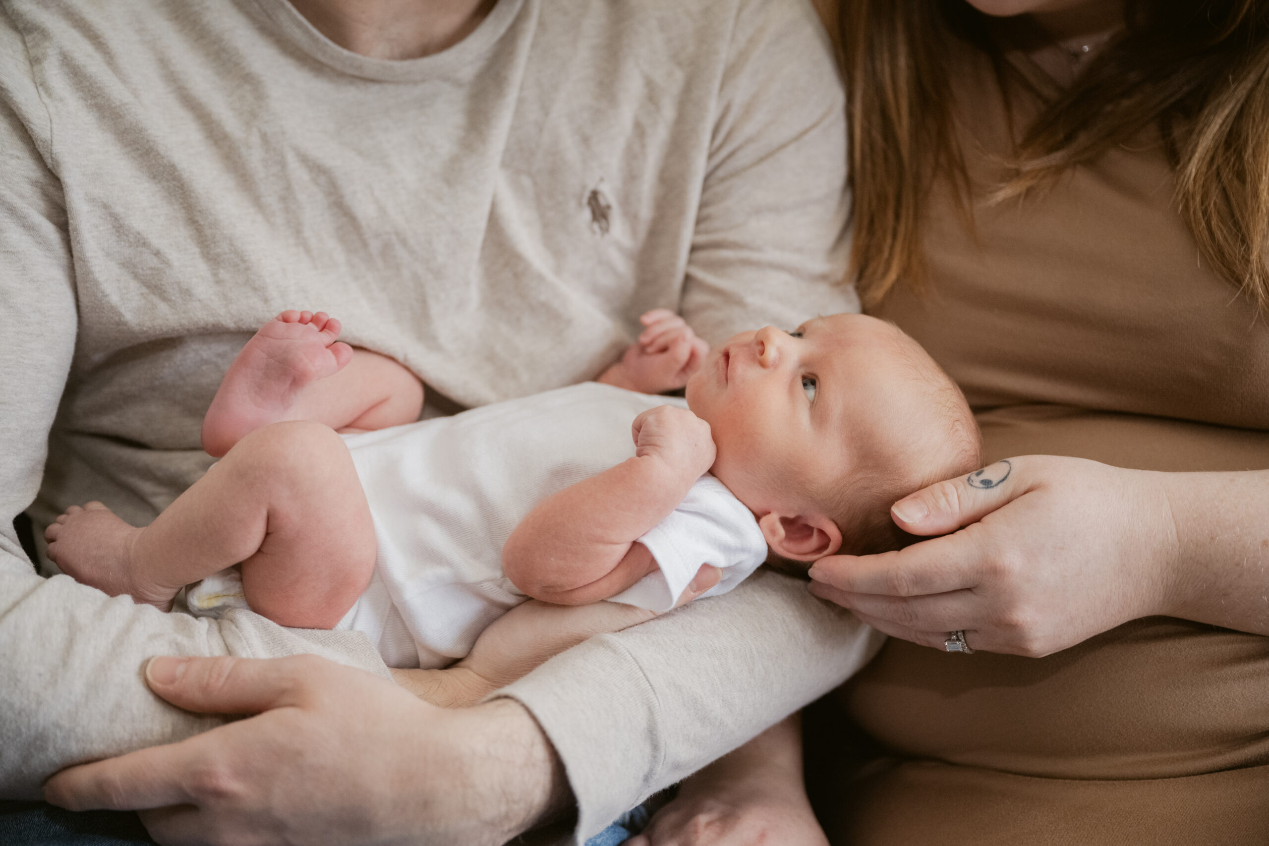 Newborn baby boy gazes up while cradled in his parents' arms during a cozy in-home photo session in Mechanicsville, VA.