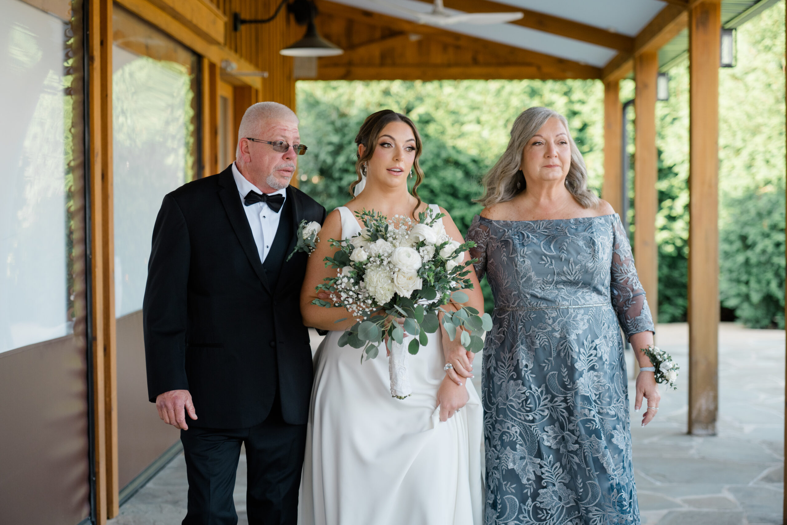 Bride walking toward the ceremony with both parents, holding a white floral bouquet at Gordon Springs in Purcellville, Virginia