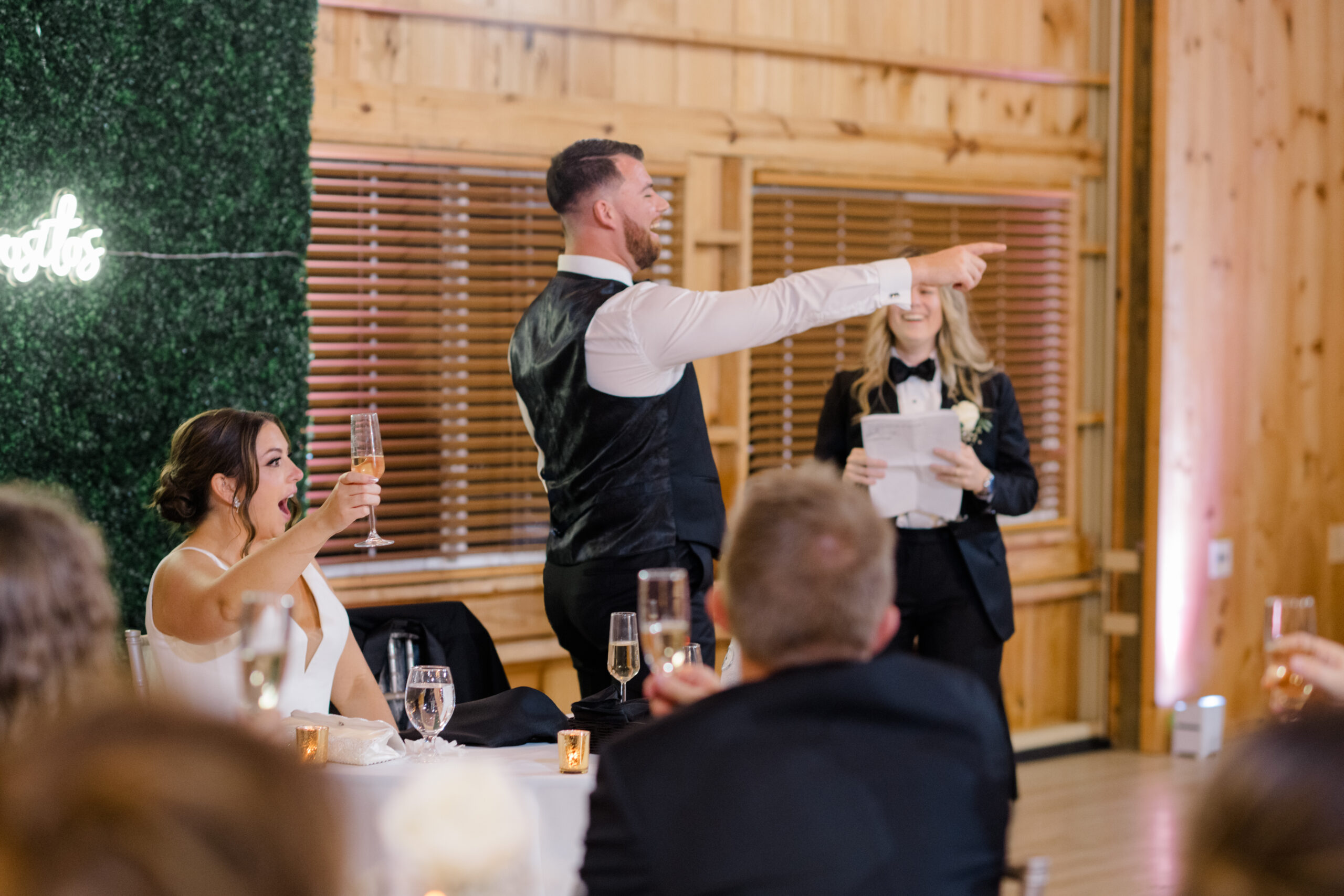 Groom raising a glass and pointing during an energetic toast at the wedding reception