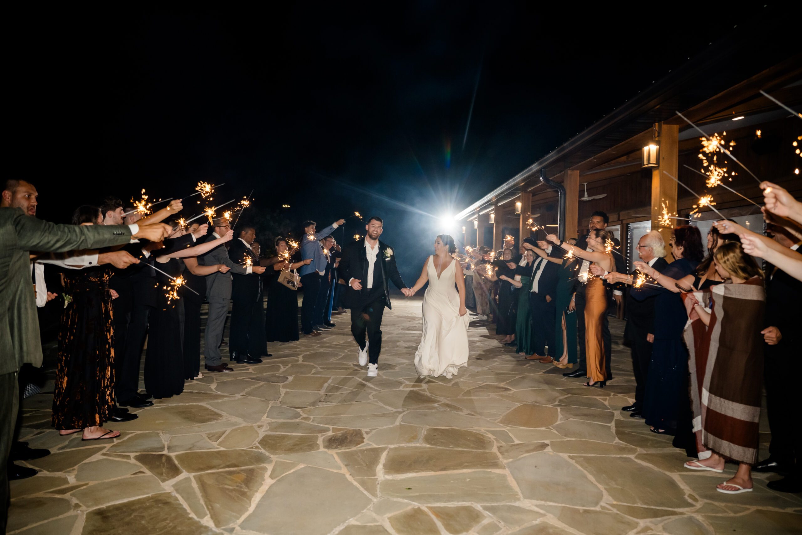 Bride and groom running hand in hand through a sparkler exit surrounded by friends and family at night