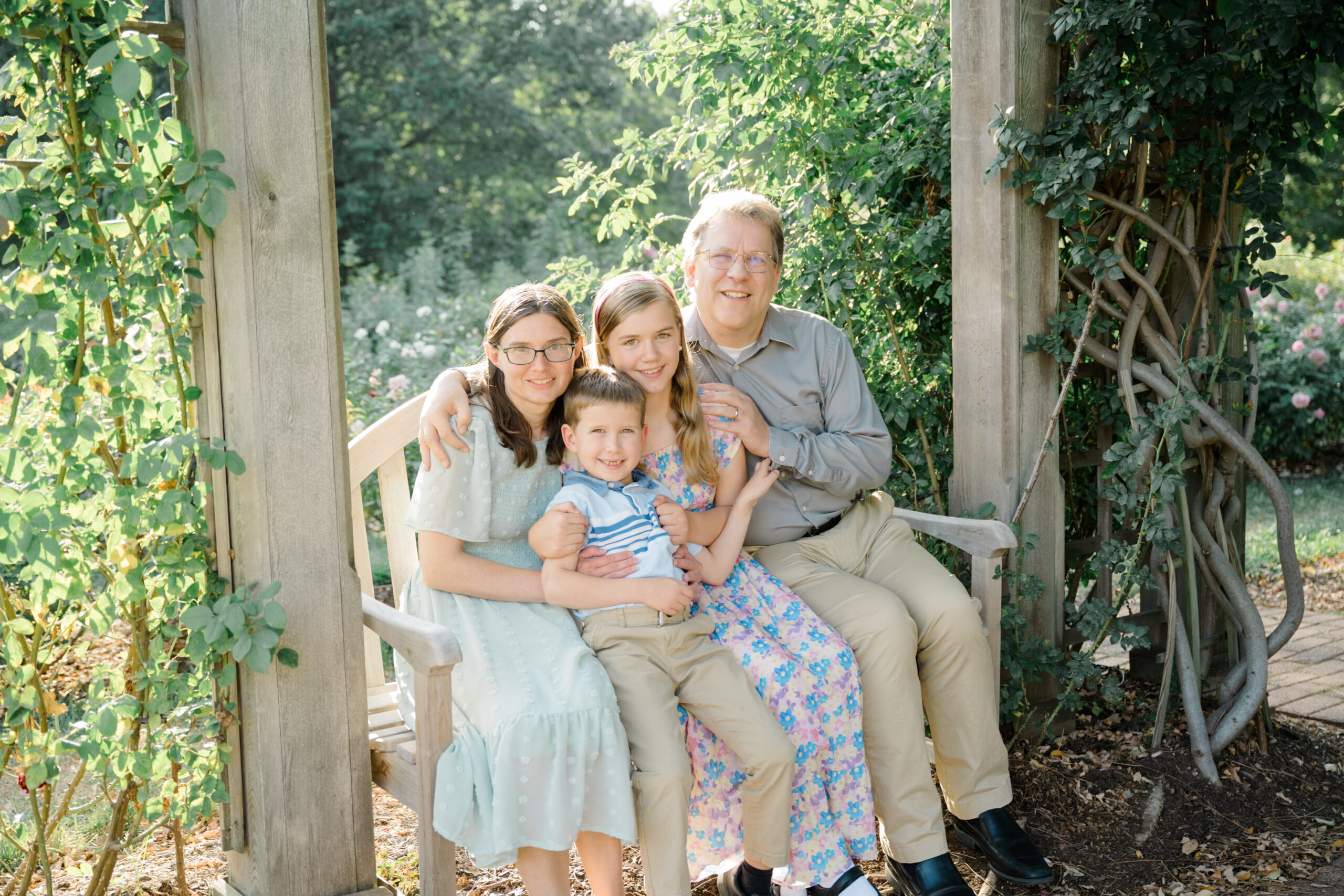 Family portrait during a golden hour mini session at the Bon Air Rose Garden, seated on a wooden bench surrounded by roses and greenery.