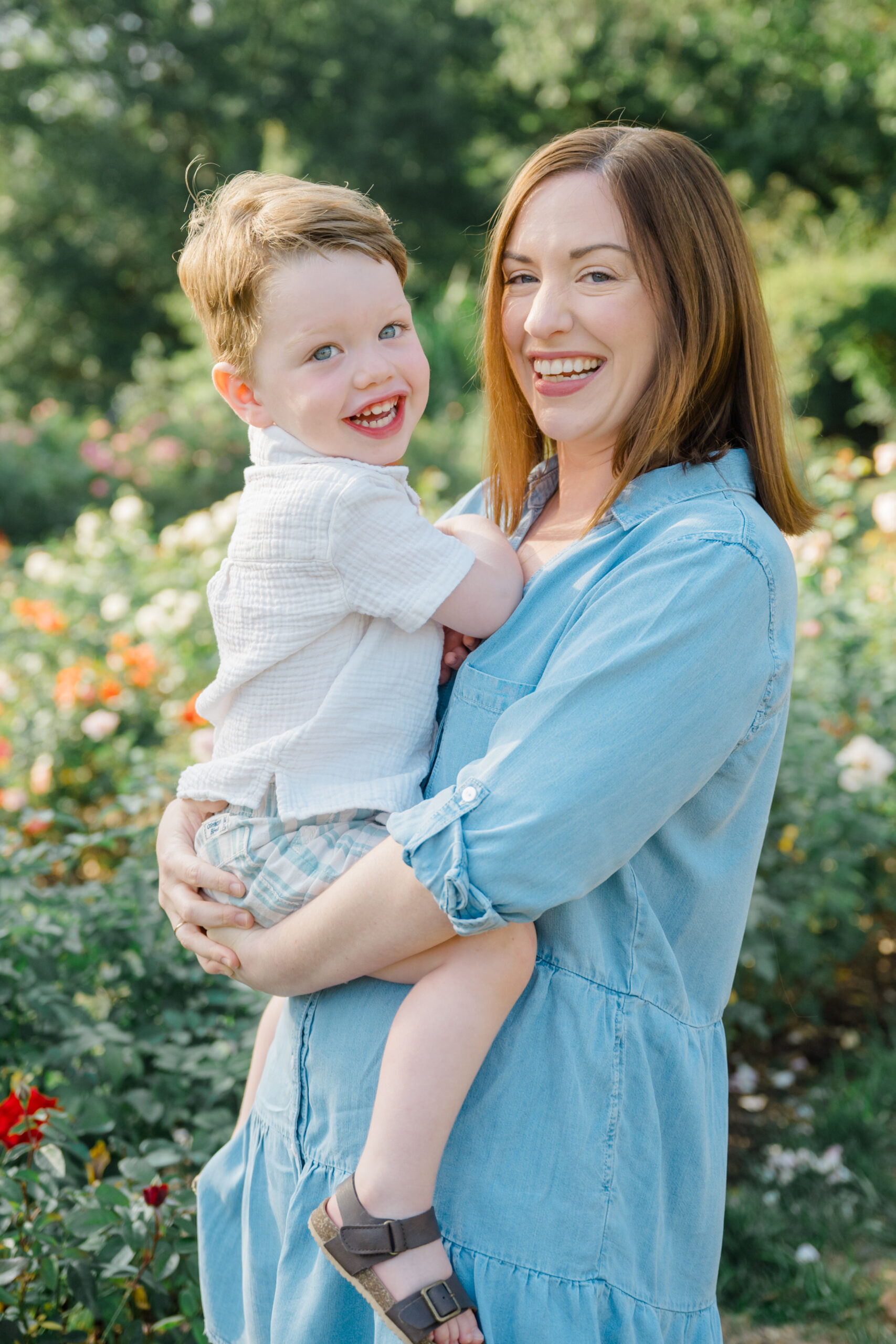 Spring family photo of a mother holding her smiling toddler in a garden filled with greenery and flowers.