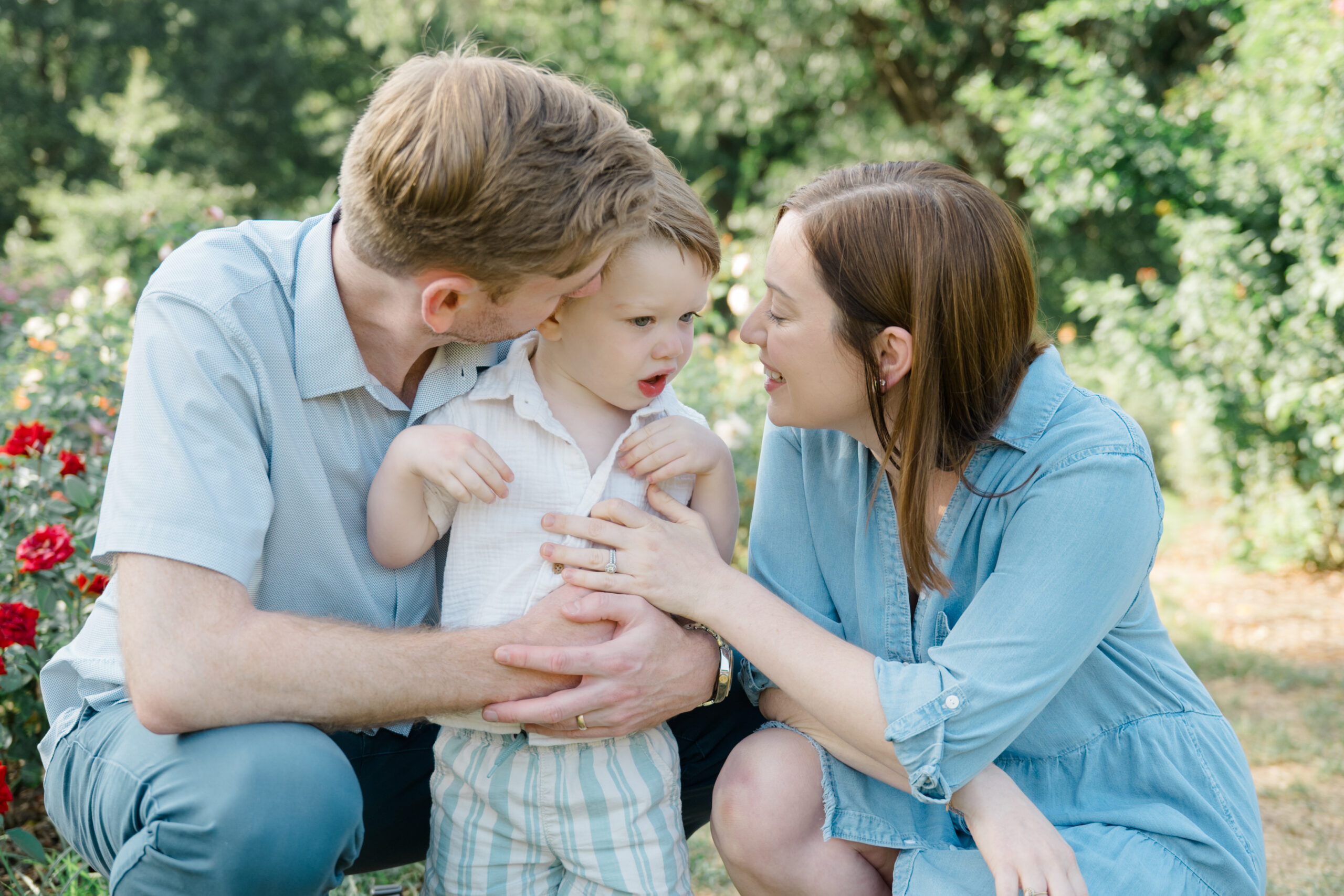 Spring family portrait of parents sitting close with their toddler, sharing a quiet moment together in a garden setting.