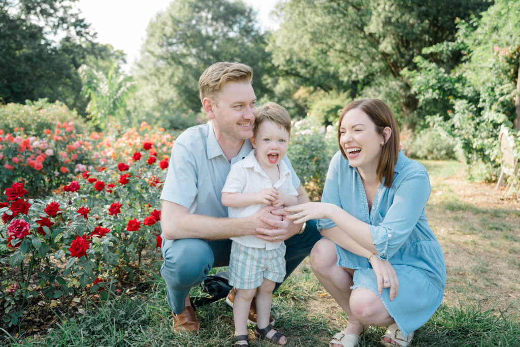 Family kneels together in the Bon Air Rose Garden during a golden hour mini session, smiling and laughing with their toddler surrounded by blooming roses.