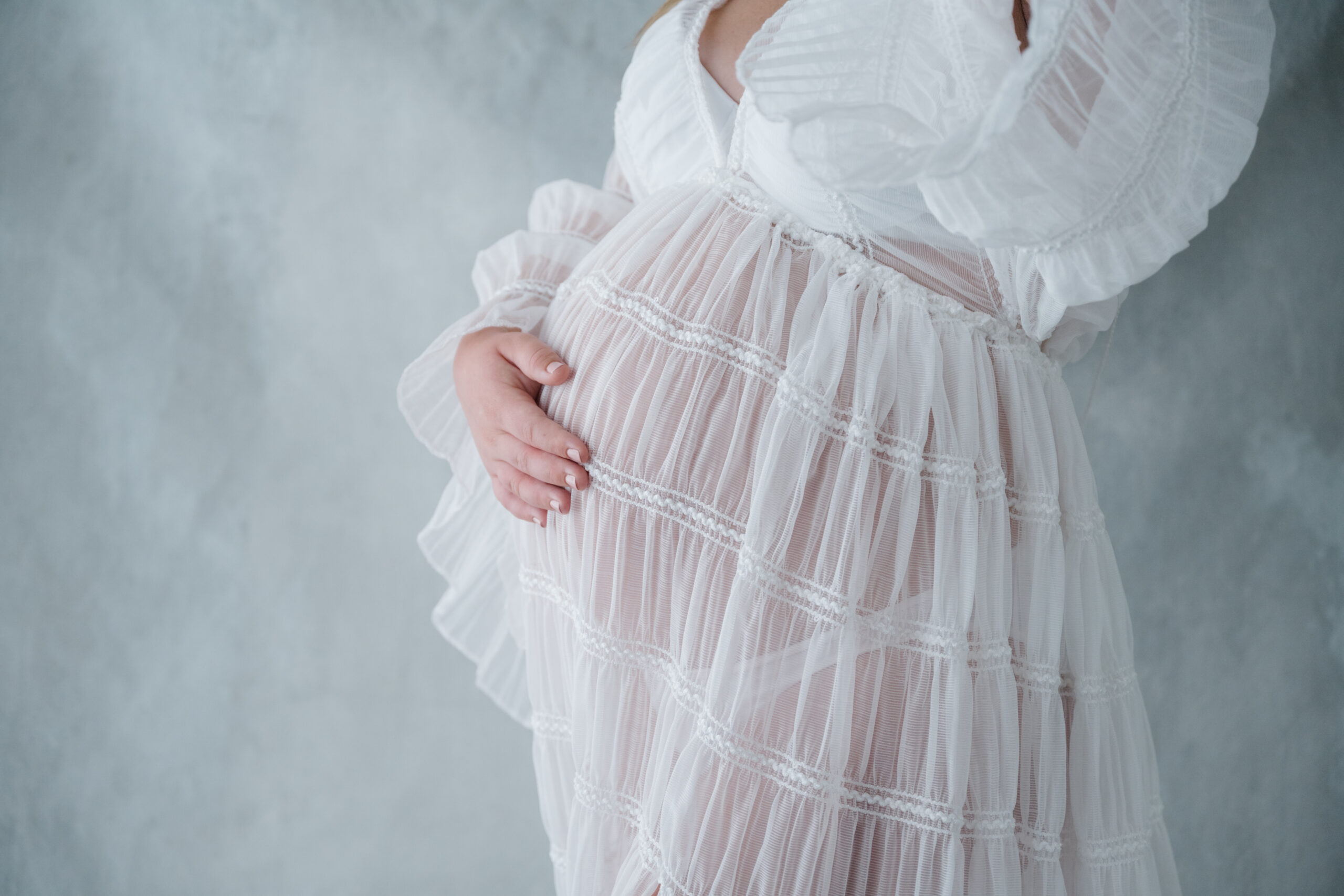 Detail maternity photo focusing on an expecting mother’s baby bump wrapped in a sheer white dress against a neutral studio backdrop.