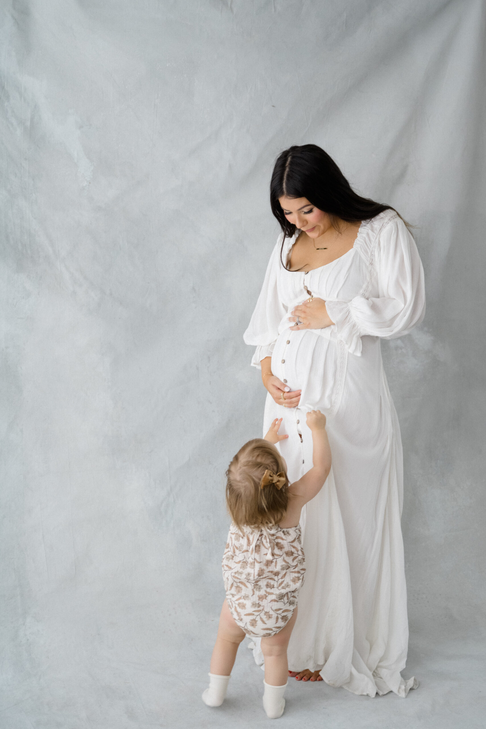 Indoor studio maternity photo of an expecting mother in a flowing white dress standing with her toddler, who is reaching up to touch her baby bump.