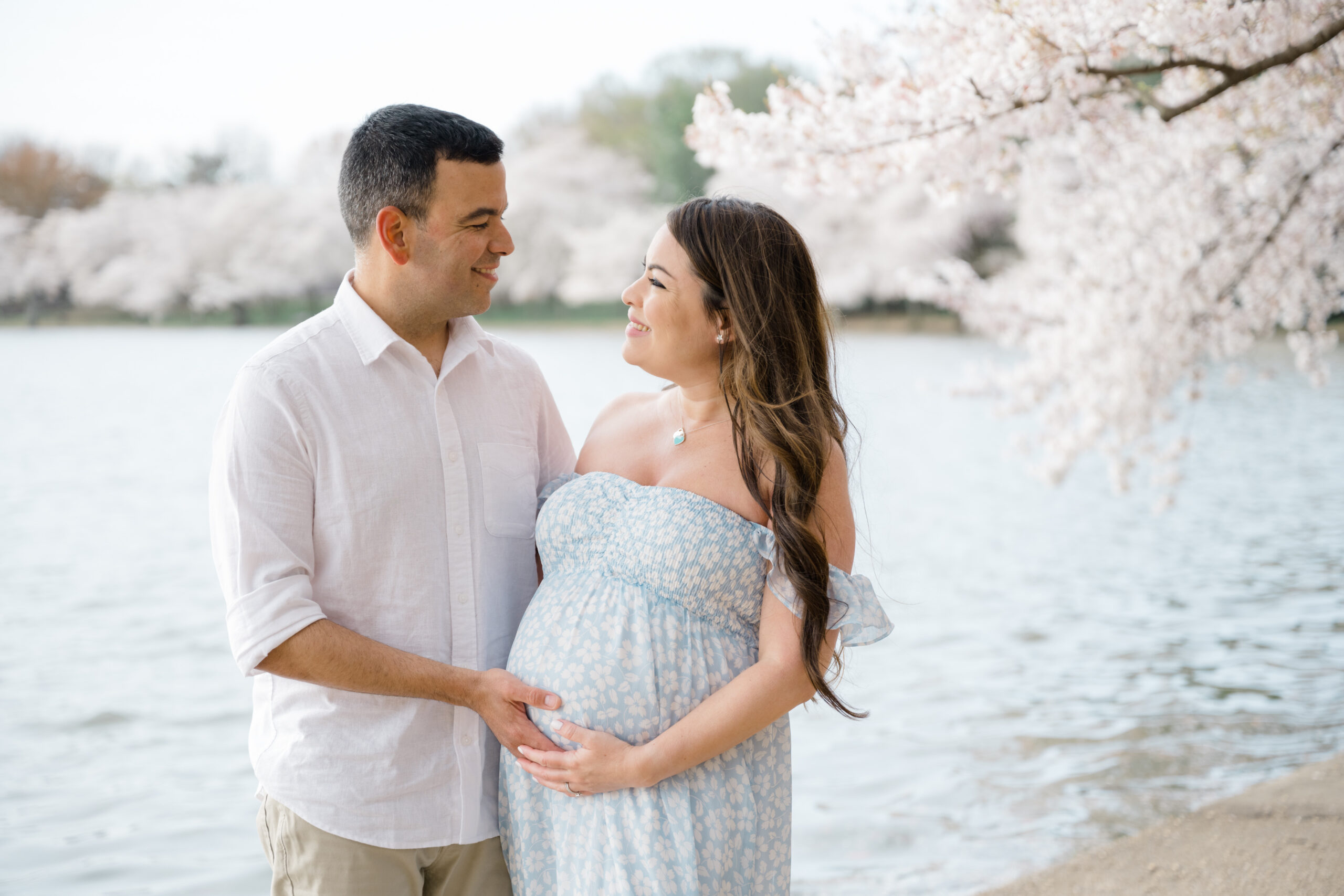 Cherry blossom maternity photo of an expecting couple standing together by the water, smiling at each other beneath blooming spring trees.