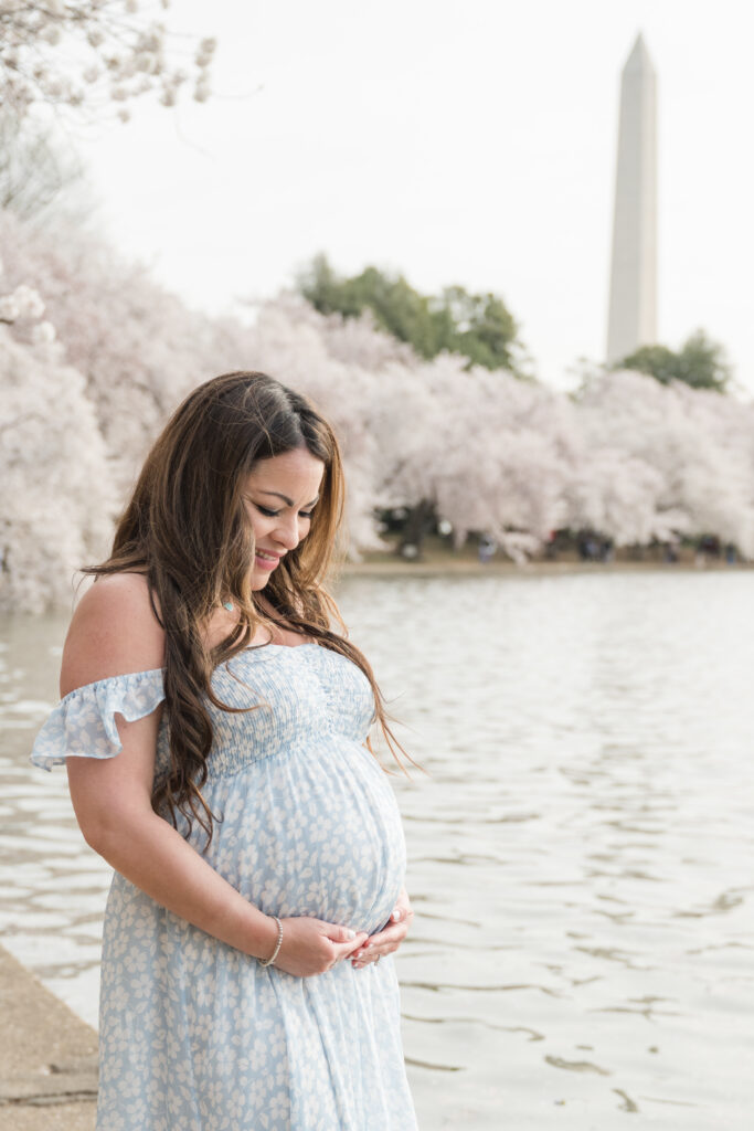 Spring maternity portrait of an expecting mother standing by the water beneath blooming cherry blossom trees, with the Washington Monument visible in the distance.