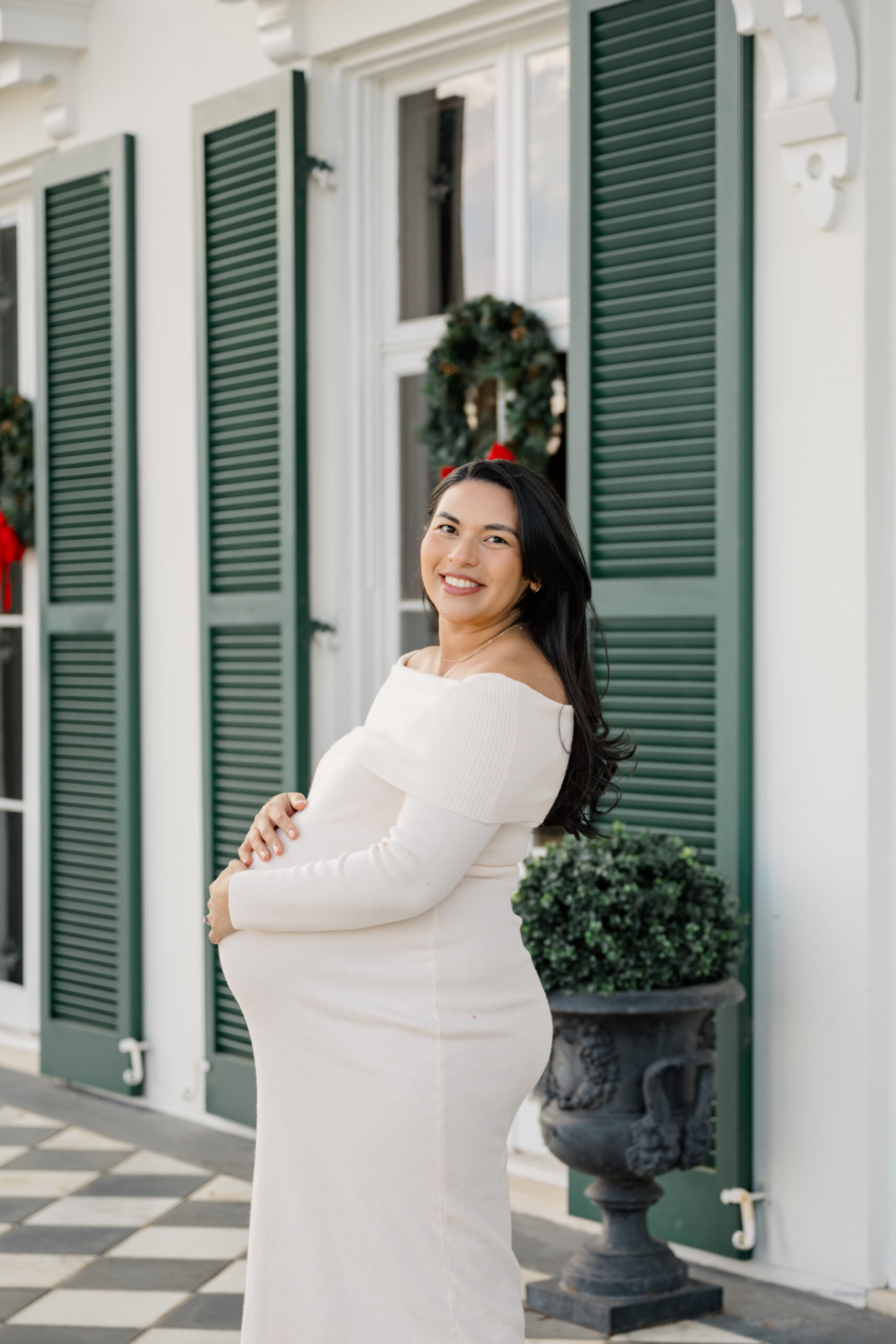 Winter maternity portrait of an expecting mother in a cream dress standing in front of a historic white home with green shutters at Morven Park.