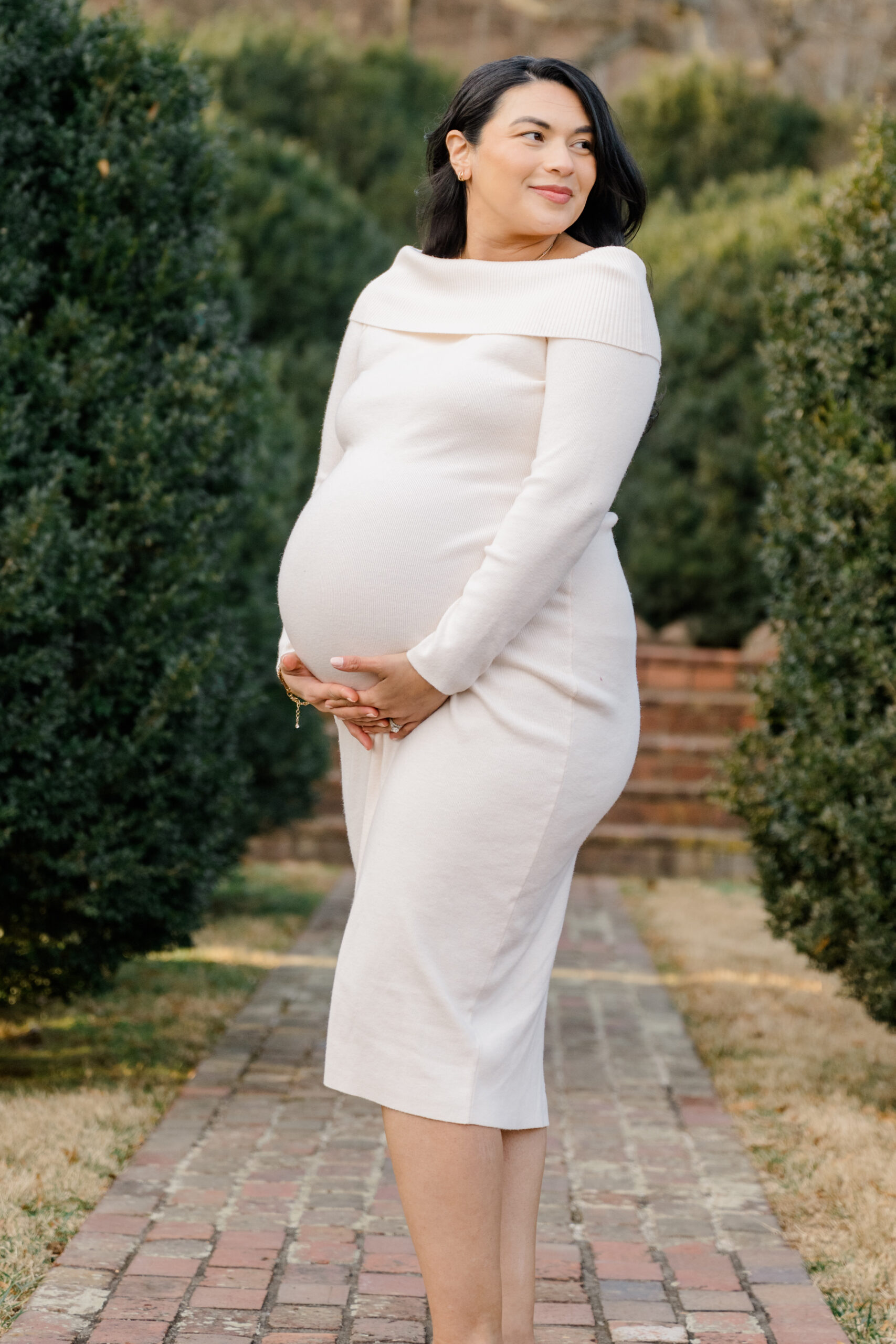 Outdoor winter maternity photo of an expecting mother in a long sleeve cream dress standing along a brick pathway surrounded by evergreen bushes.