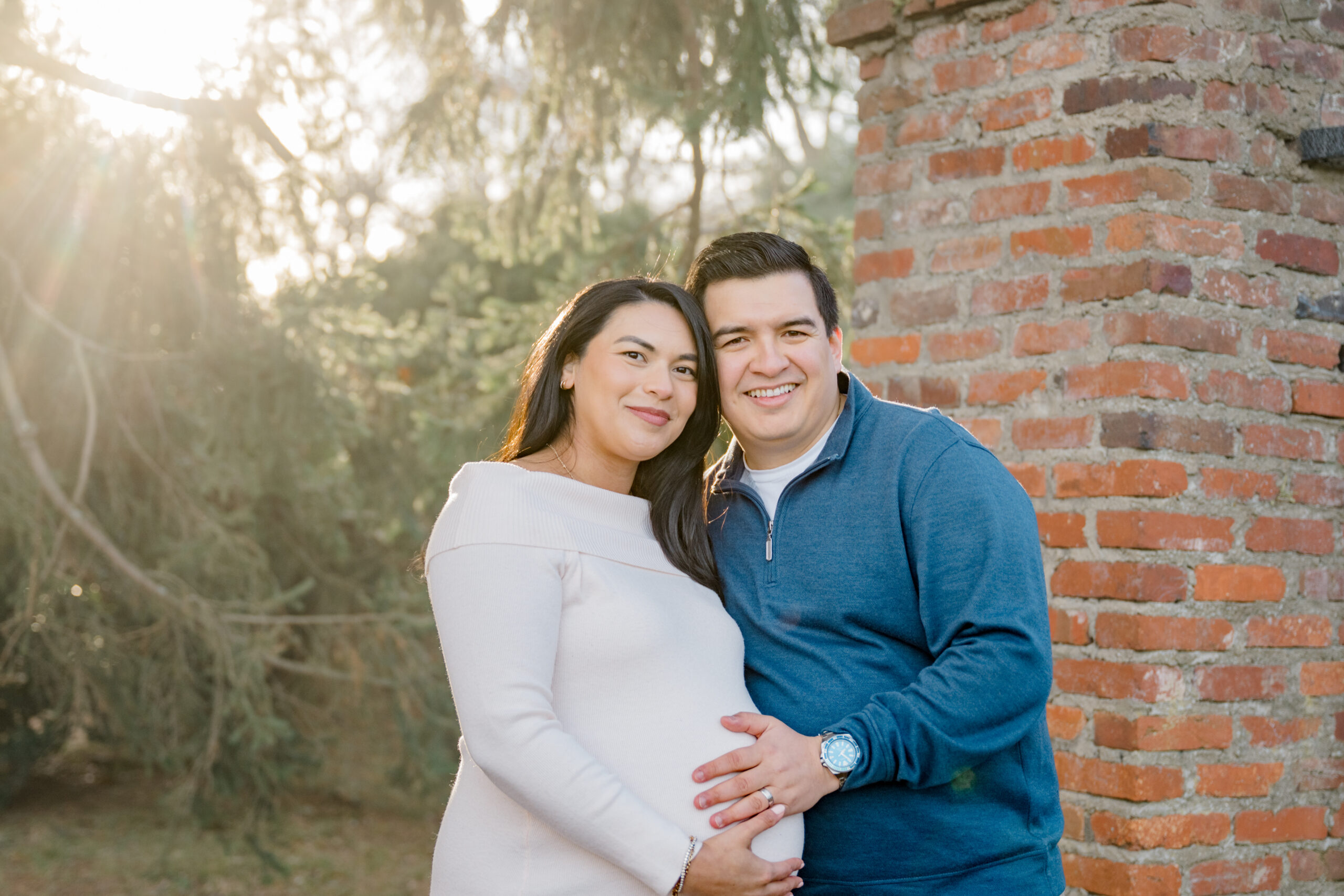 Golden hour winter maternity photo of an expecting couple standing together near a brick wall with soft sunlight filtering through evergreen trees.