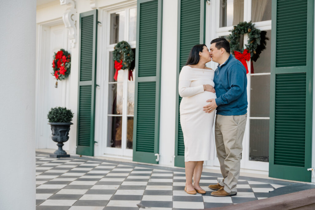 Winter maternity portrait of an expecting couple standing together on the porch at Morven Park, decorated with holiday wreaths.