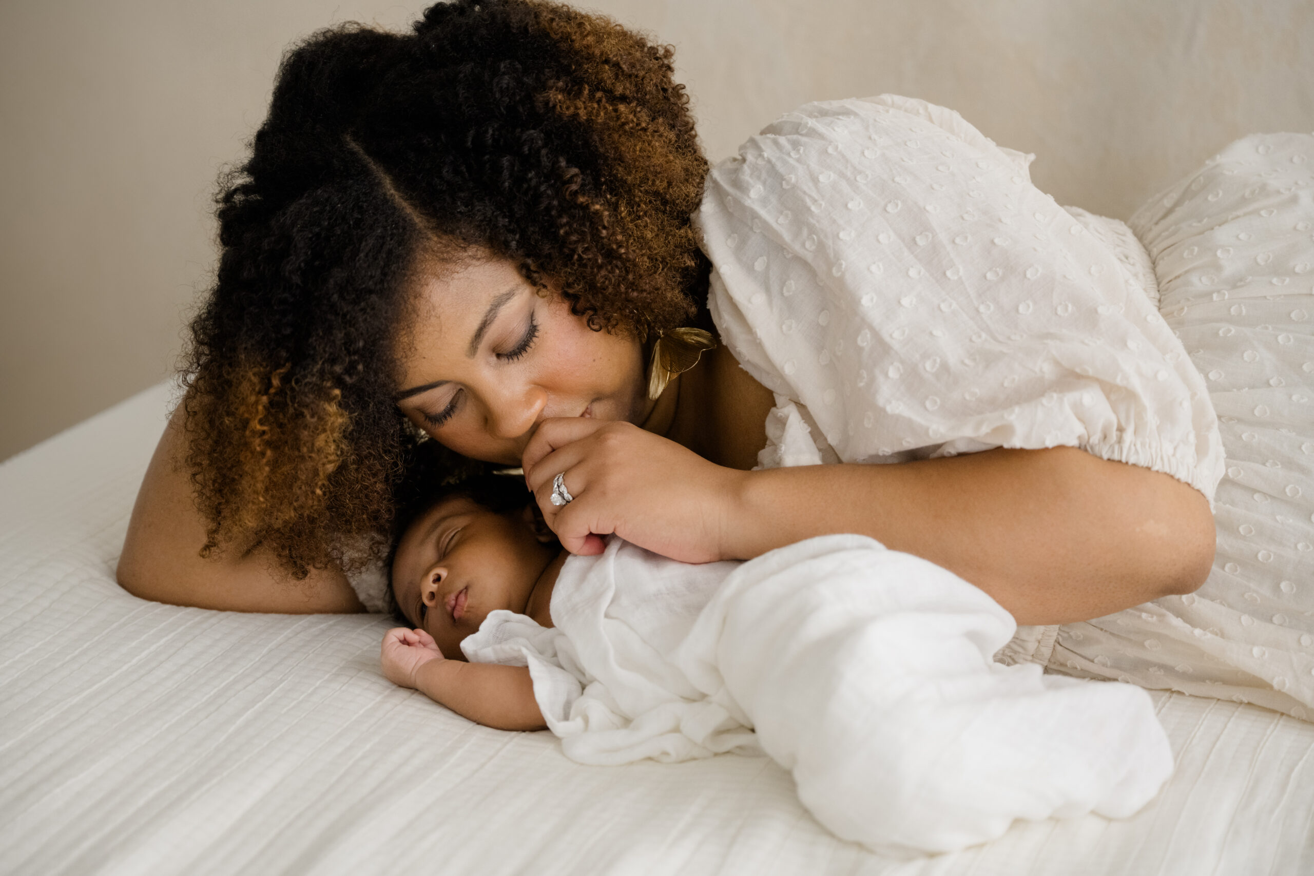 Intimate motherhood photo of a mother lying beside her sleeping newborn, gently kissing the baby’s hand.