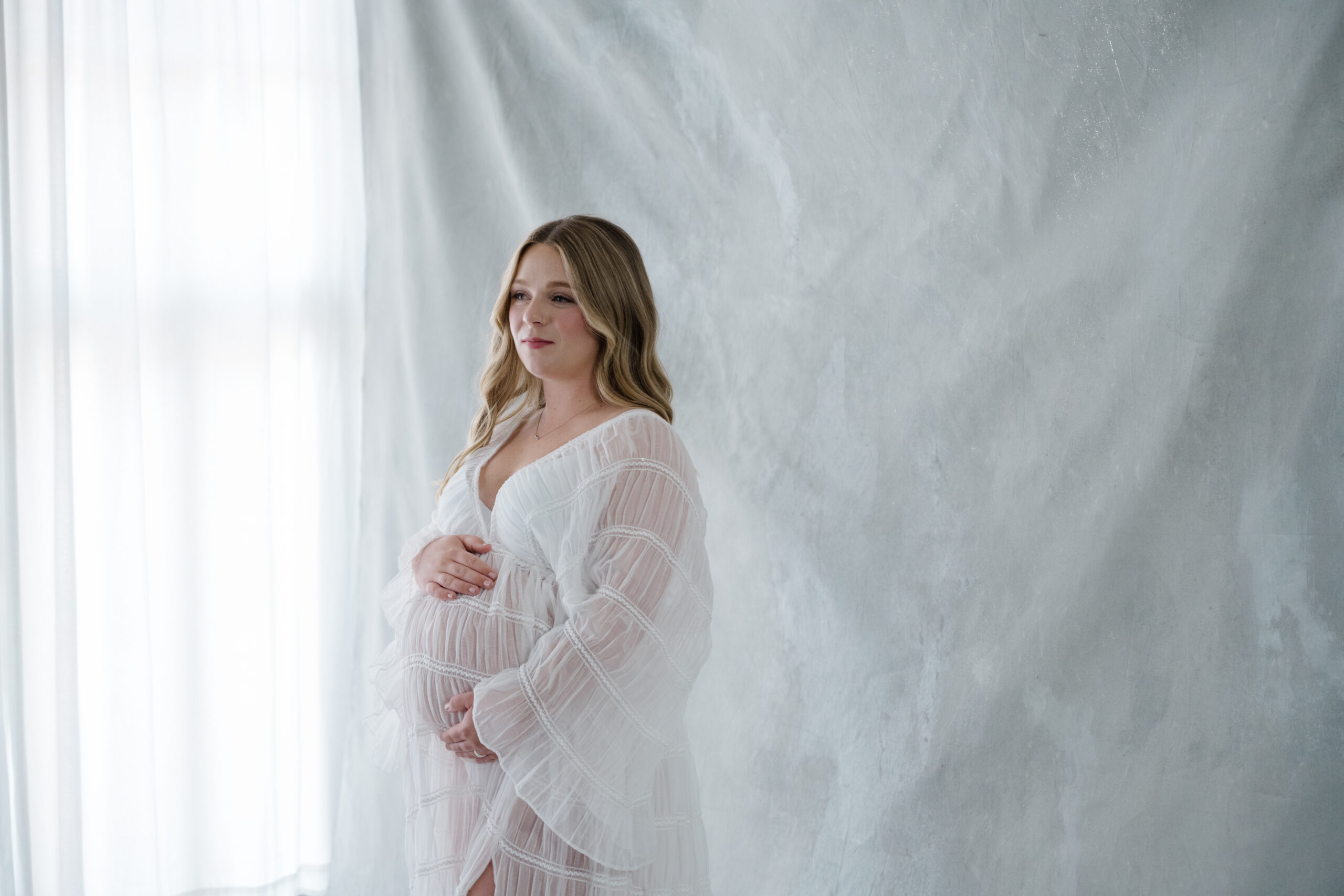 Studio maternity portrait of an expecting mother in a sheer white dress standing in soft natural light against a neutral backdrop.