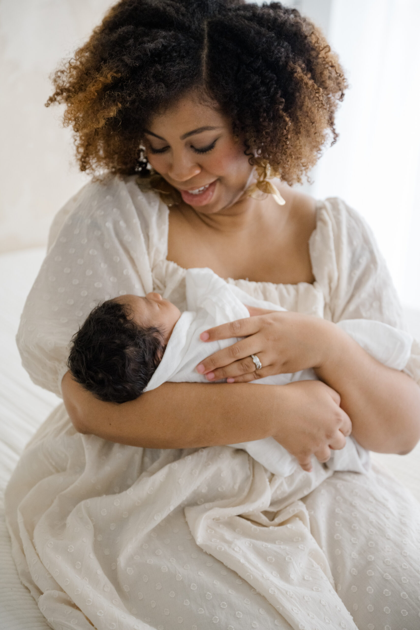 Motherhood portrait of a mother cradling her newborn in her arms while sitting indoors in soft window light.