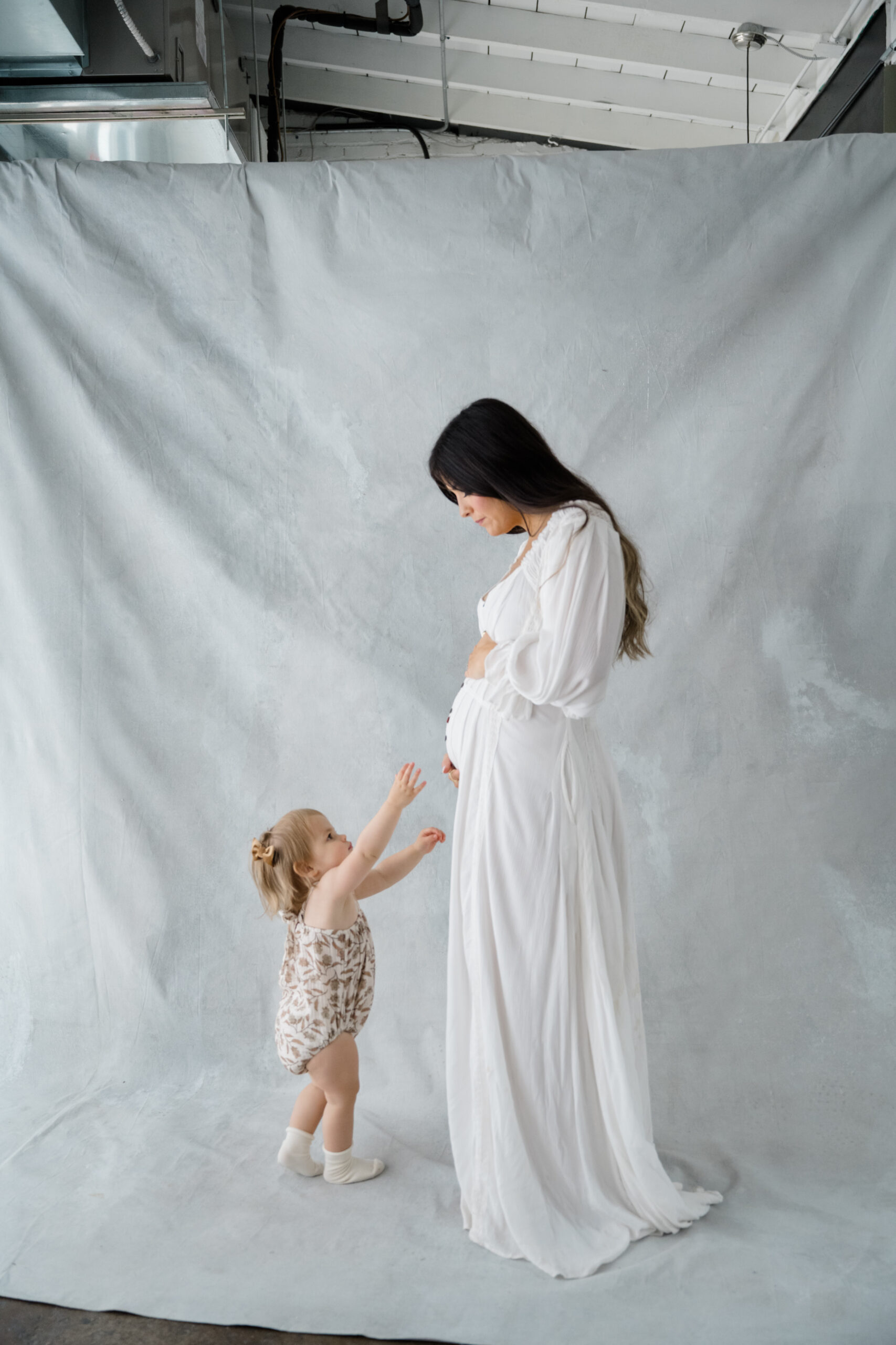 Wide studio maternity portrait of an expecting mother and her toddler standing together against a soft neutral backdrop.